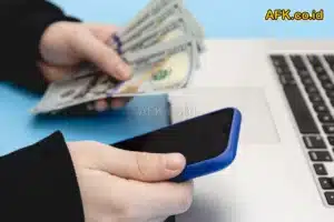 Tangan laki-laki yang memakai jas, memegang hp dan uang di atas meja, dengan laptop
