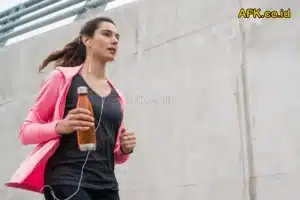 perempuan yang sedang jogging menuruni tangga dengan jaket pink dan membawa botol minum serta memakai handsfree