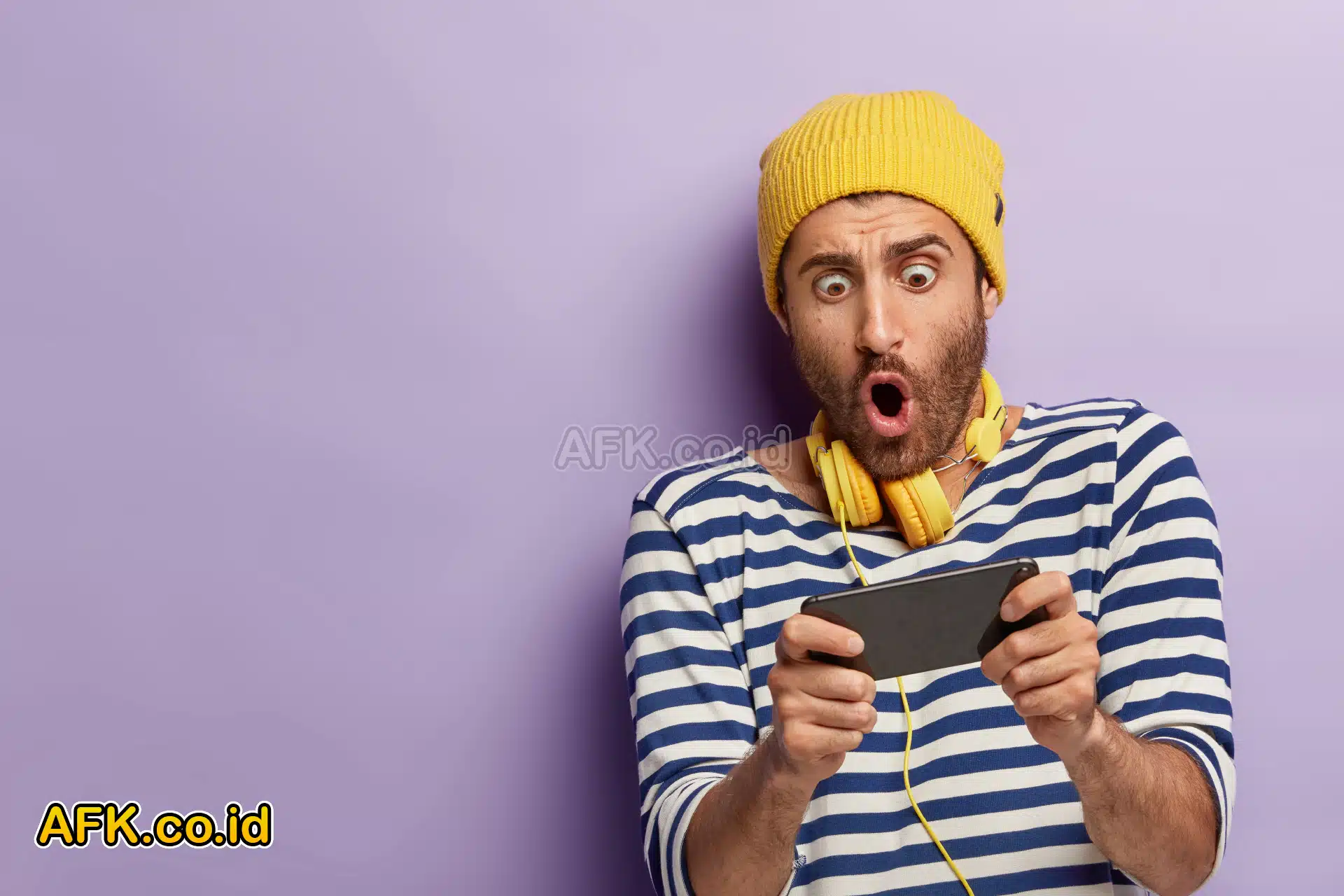 Laki-laki berjenggot tipis memegang handphone dengan ekspresi terkejut memakai topi kuning dan headset kuning